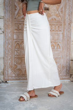 - Product: A white linen skirt with a long, maxi-length silhouette and a subtle front drape.
- Intended audience: Women (judging from the cut, fit, and styling).
- Notable attributes: White linen fabric, high waist, wrap-like front detail, full-length / maxi style, casual slide sandals, earthy-toned backdrop.